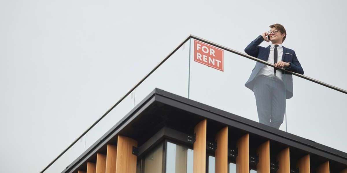 Wide angle view at smiling real estate agent speaking by phone while standing on roof of modern building next to red FOR RENT sign