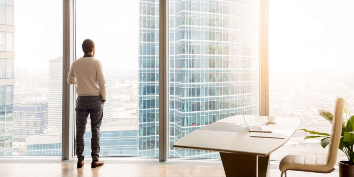 Rear view at young successful businessman standing in office looking through full-length window at cityscape with skyscrapers