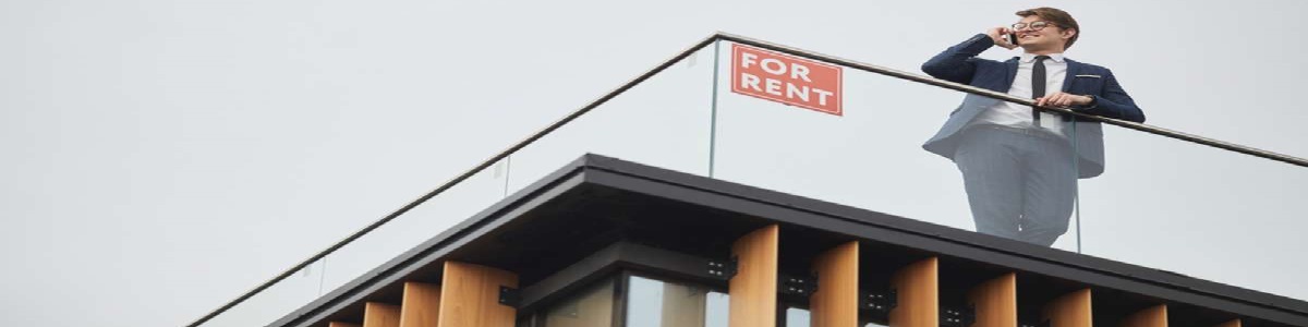 Wide angle view at smiling real estate agent speaking by phone while standing on roof of modern building next to red FOR RENT sign